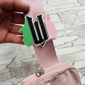 May include: A pink crossbody bag with a matching strap. A decorative emblem with the word "Lil" in black and green and pink profile silhouettes is attached to the strap. The text "Dixieland's Design Co." is also visible.