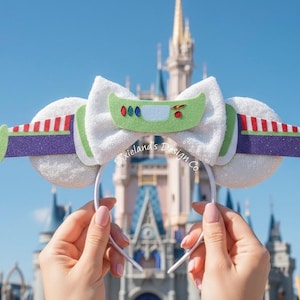 May include: White, glittery mouse ears with a Buzz Lightyear theme. The ears have a white bow with a green and red design, and purple and red accents. The background features a castle.