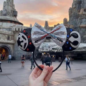 May include: Black and silver sequined mouse ears with a silver bow featuring red and blue lightsaber accents. The ears have a black Imperial symbol and a silver Rebel Alliance symbol. The headband is black.