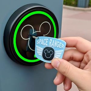Może przedstawiać: Jasnoniebieski brelok Space Ranger z logo Myszki Miki i skrzydłami jest trzymany w pobliżu czarnego czytnika kołowego z zieloną obwódką i logo Myszki Miki. Czytnik znajduje się na szarej powierzchni.