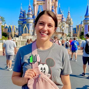 May include: A woman smiles in front of a fairytale castle. She wears a gray t-shirt with a Mickey Mouse graphic and a pink fanny pack. The background shows a sunny day with other people walking around.