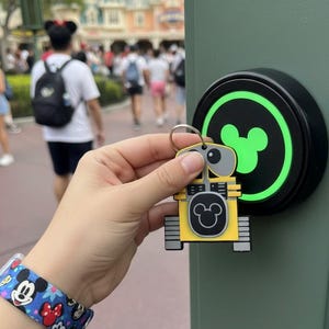 May include: A yellow and gray WALL-E keychain with a Mickey Mouse icon on the front. The keychain is held near a green-lit button with a Mickey Mouse icon. A Mickey Mouse bracelet is visible on the wrist.