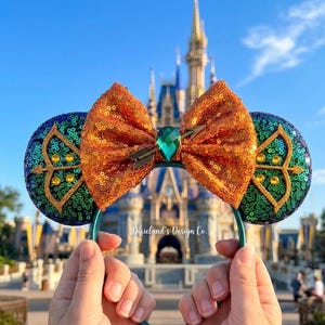 Op de afbeelding: Een paar teal muisoren met een grote oranje pailletten strik en een groene edelsteen. De oren hebben een gouden pijl-en-boog ontwerp. De achtergrond toont een kasteel en een blauwe lucht.