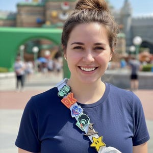 May include: A woman wearing a navy blue t-shirt and a white fanny pack. A decorative strap across her chest features colorful cartoon character pins, including a yellow star and the letters "DK". The background includes a blurred amusement park.