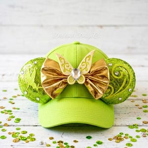 May include: Lime green baseball cap with gold sequined ears and a gold bow with a central jewel. The bow has a decorative design. The ears are embellished with sequins and a silhouette. Scattered sequins add a touch of sparkle.