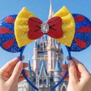 Pode incluir: Um par de orelhas de rato com o tema Branca de Neve. As orelhas são azuis com detalhes de lantejoulas vermelhas, um laço de lantejoulas amarelo e um laço vermelho com um centro de joia. A tiara é azul.