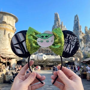 May include: Black headband with mouse ears. The ears feature a green sequined bow and a Baby Yoda graphic. One ear has a black and white design, the other ear has the text "THIS IS THE WAY" in white.