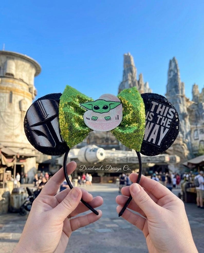 May include: Black headband with mouse ears. The ears feature a green sequined bow and a Baby Yoda graphic. One ear has a black and white design, the other ear has the text "THIS IS THE WAY" in white.