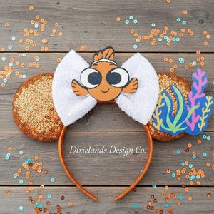 Peut inclure: Un serre-tête orange avec des oreilles à paillettes, un nœud blanc et un motif de poisson Nemo. Le serre-tête comporte également un accent de corail et d'algues. Le design est posé sur un fond en bois avec des paillettes orange et bleues éparpillées.
