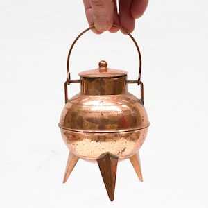 Puede incluir: Un pequeño caldero de cobre vintage con tapa y asa. El caldero tiene tres patas puntiagudas y un cuerpo redondo. El cobre tiene un tono marrón rojizo cálido, y el asa se arquea sobre la parte superior. El caldero es sostenido por una mano.