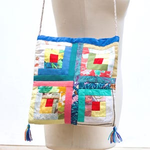 May include: A square crossbody bag constructed from a patchwork of various colourful fabric scraps in shades of red, yellow, green, and blue. The bag features a blue top border, a thin patterned strap, and two small multi-coloured yarn tassels at the bottom corners.