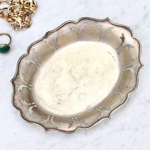 Puede incluir: Bandeja decorativa ovalada plateada con borde festoneado y diseños calados ornamentados. Una cadena dorada y dos anillos son visibles en el fondo. La bandeja es ideal para exhibir joyas o pequeños objetos.