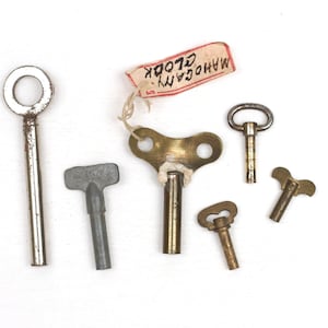 May include: A collection of five metal clock keys of varying sizes and designs, including silver-toned and brass-toned finishes. One brass key features a white string attached to a rectangular paper tag with the handwritten text MAHOGANY CLOCK.