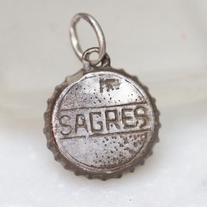 May include: Silver charm shaped like a bottle cap with the word "SAGRES" embossed on it.