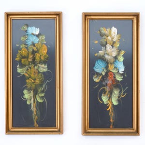 May include: Two rectangular wall art pieces in gold frames, each featuring a vertical floral arrangement painted on a black background. The flowers are rendered in shades of blue, white, yellow, and green with decorative swirling brushstrokes.