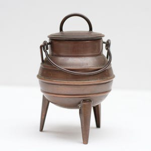 Op de afbeelding: Een kleine, antieke koperen ketel met een deksel en een handvat. De ketel heeft drie poten en een handvat op het deksel. Het koper heeft een warme, roodbruine kleur. De ketel wordt waarschijnlijk gebruikt om te koken of voor decoratieve doeleinden.