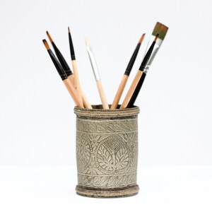 May include: A cylindrical metal container with an aged silver finish and embossed leaf patterns, holding a collection of various paintbrushes with wooden handles and black or silver ferrules, featuring both fine-tipped and flat-bristled brushes.