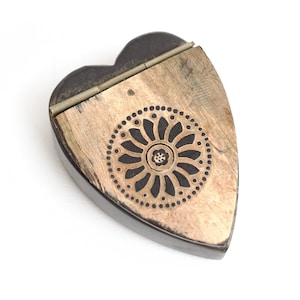 Puede incluir: Una caja en forma de corazón con base de color marrón oscuro y tapa abatible. La tapa presenta una incrustación texturizada de color claro con un motivo circular negro central que contiene una flor estilizada rodeada de puntos.
