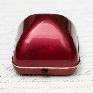 May include: A red ring box with a gold trim. The box is closed and has a slight curve to the top.