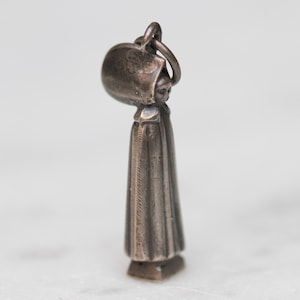 May include: A small, antique silver charm in the shape of a woman wearing a bonnet and long dress. The charm has a loop at the top for attaching to a bracelet or necklace. The woman's face is detailed.
