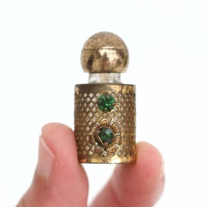 May include: A miniature perfume bottle featuring a cylindrical gold-toned metal casing with a diamond-patterned lattice overlay. The casing is decorated with two small, faceted green glass stones, one set within a floral-shaped metal mount.