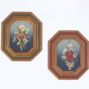 May include: Two octagonal wall frames with gold-toned inner borders. The left frame has a dark green outer edge and a painting of pink and orange flowers. The right frame has a dark red outer edge and a painting of blue and red flowers.