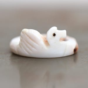 Può includere: Piccola statuina di cigno bianco scolpito, con un'ala dettagliata e un piccolo occhio scuro. Il cigno è appoggiato su una base circolare e l'intaglio ha un aspetto leggermente invecchiato.