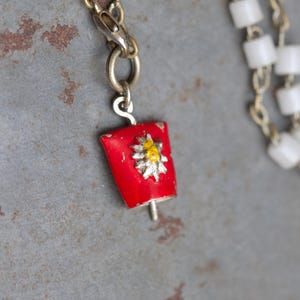 Peut inclure: Un pendentif vintage en forme de cloche en émail rouge, orné d'un motif floral jaune et argenté. Le pendentif est suspendu à une chaîne argentée, avec un collier de perles blanches visible en arrière-plan. Un bijou unique.