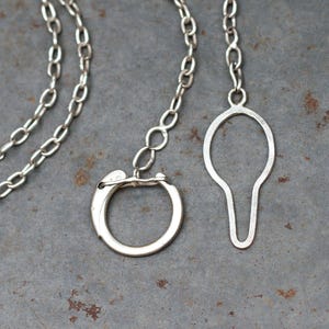 May include: A silver-colored metal chain necklace featuring a circular clasp and a separate teardrop-shaped pendant. The necklace consists of oval-shaped links connected to a round ring closure, with a distinct elongated bulbous charm attached.
