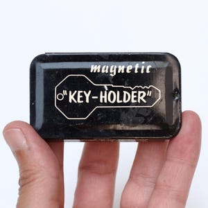 May include: A small, rectangular black metal box with rounded corners. The lid features the white text "magnetic" above a white outline graphic of a key containing the text "KEY-HOLDER" in a stylized font. The surface shows minor wear.