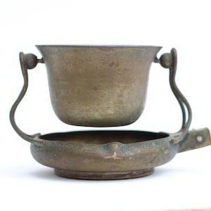 Op de afbeelding: Een vintage koperen vat met een diepe, ronde kom opgehangen tussen twee gebogen metalen armen die vastzitten aan een brede, ondiepe ronde basis. Het metalen oppervlak vertoont een verweerde, verouderde patina met zichtbare donkere vlekken.