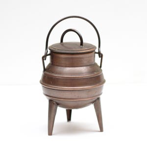 Può includere: Un piccolo calderone antico in rame con coperchio e manico. Il calderone ha tre gambe e un manico curvo per il trasporto. Il rame ha una calda tonalità marrone-rossastra e il design suggerisce un'estetica vintage o rustica.