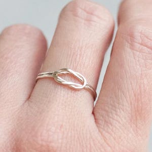 Op de afbeelding: Een zilverkleurige ring met een knoopontwerp. De ring wordt aan een vinger gedragen, waarbij de ingewikkelde knoopdetails worden getoond. De band van de ring is dun en de knoop is het middelpunt van het sieraad.