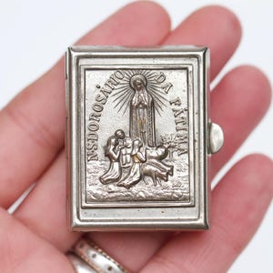 Pode incluir: Uma caixa metálica retangular de tom prateado com um design em relevo da Virgem Maria aparecendo a três crianças. O texto "N.S. DO ROSÁRIO DA FÁTIMA" está gravado em torno da figura central. A caixa possui um fecho lateral articulado.