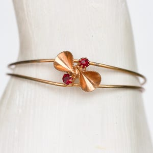 Puede incluir: Una pulsera rígida de metal dorado con un elemento decorativo central en forma de dos hojas en abanico y dos pequeñas piedras rojas redondas y facetadas engastadas. La pulsera consta de dos bandas metálicas doradas finas y paralelas.