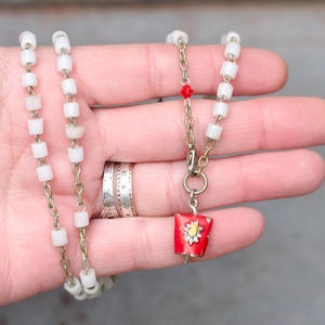 Peut inclure: Un collier vintage composé de perles blanches rectangulaires enfilées sur une chaîne dorée. Le collier comporte un pendentif rouge avec un motif floral jaune et blanc. Le pendentif est rectangulaire et possède une petite boucle.
