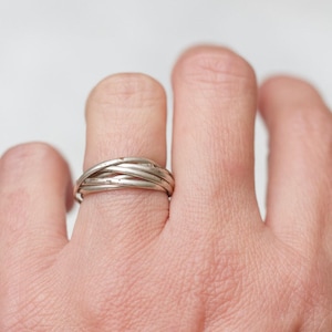 May include: A silver ring with a woven design is worn on a finger. The ring appears to be made of multiple strands of metal intertwined to create a textured, band-style ring. The background is a soft, neutral color.
