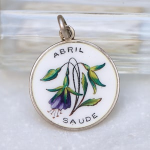 May include: A silver charm with a white enamel background featuring a purple flower with green leaves. The charm is inscribed with the words "ABRIL" and "SAUDE".