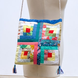 Op de afbeelding: Een kleurrijke patchwork crossbody tas met een vierkant ontwerp. De tas heeft een verscheidenheid aan stoffen in tinten blauw, groen, geel, rood en roze, gerangschikt in een blokhutpatroon. Het heeft een blauwe rand, een geweven band en kwastjes aan de onderkant.