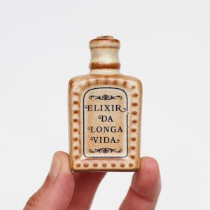 May include: A small, rectangular ceramic bottle with a tan, speckled glaze and a narrow neck. The front features a decorative black border and the printed text ELIXIR DA LONGA VIDA in a serif font, surrounded by ornate scrollwork details.