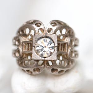 May include: A silver ring with a central square setting holding a clear, faceted gemstone. The ring has an openwork design with circular and rectangular cutouts, creating a delicate, vintage aesthetic.