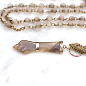May include: A necklace featuring a series of translucent, frosted grey beads strung on a gold-toned metal chain. The focal point is a carved grey stone pendant shaped like a hand, accented with a metallic band and a textured, leaf-shaped toggle clasp.