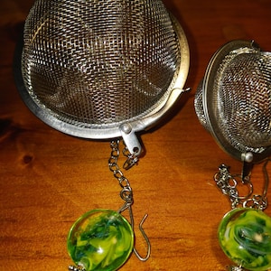 May include: Two stainless steel tea infusers with a mesh basket design. Each infuser has a chain attached to a green and yellow glass bead. The infusers are resting on a wooden surface.