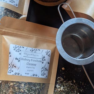 May include: A package of "Kidney Function Cleanse" organic tea from PhytoPharm, with a metal tea infuser and a brown mug. The tea package is in a brown paper bag, and the tea is visible inside. The image is taken from above.