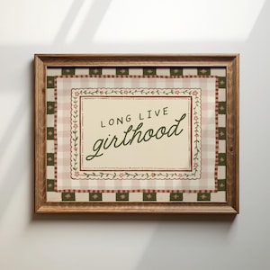 Puede incluir: Un estampado enmarcado con las palabras "LONG LIVE girlhood" en una fuente decorativa. El estampado tiene un fondo color crema con un patrón floral y de cuadros en tonos rosa, verde y marrón. El marco es de madera.