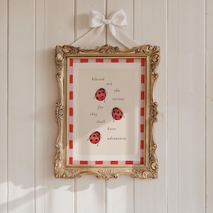 Könnte beinhalten: Ein gerahmter Druck mit einem goldenen, verzierten Rahmen, der den Text "blessed are the curious for they shall have adventures" und drei Marienkäfer-Illustrationen zeigt. Der Rahmen ist mit einer weißen Schleife verziert.