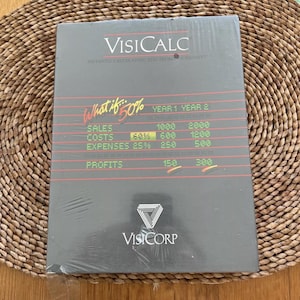 May include: A sealed VisiCalc software package. The cover is black with red and white text, including "VisiCalc" and "What if 50%" with financial data. The VisiCorp logo is at the bottom.