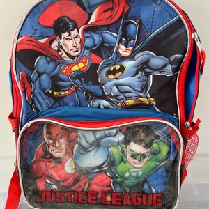 May include: A blue backpack featuring Superman and Batman on the top panel, with The Flash and Green Lantern below. The backpack has red trim, a red shoulder strap, and the words "JUSTICE LEAGUE" across the bottom.