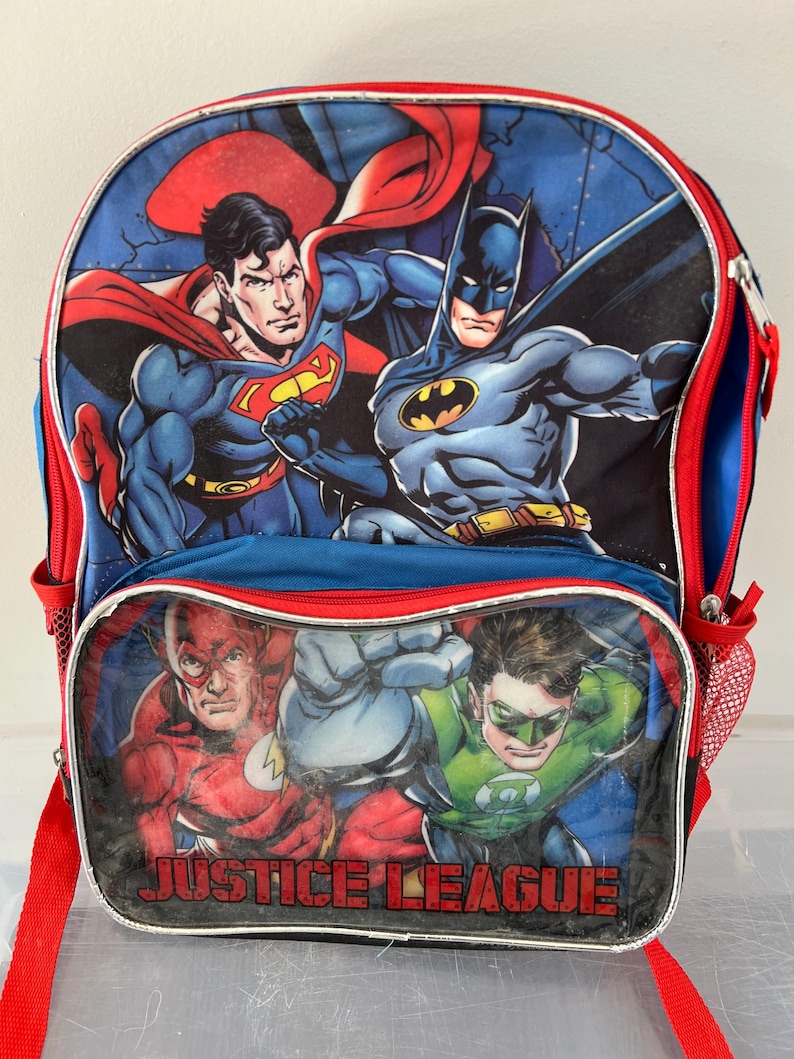 May include: A blue backpack featuring Superman and Batman on the top panel, with The Flash and Green Lantern below. The backpack has red trim, a red shoulder strap, and the words "JUSTICE LEAGUE" across the bottom.