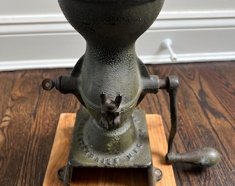 Puede incluir: Un antiguo molinillo de café Enterprise con acabado verde oscuro. El molinillo tiene una tolva, una manivela y está montado sobre una base de madera. La base tiene el texto "ENTERPRISE MFG".